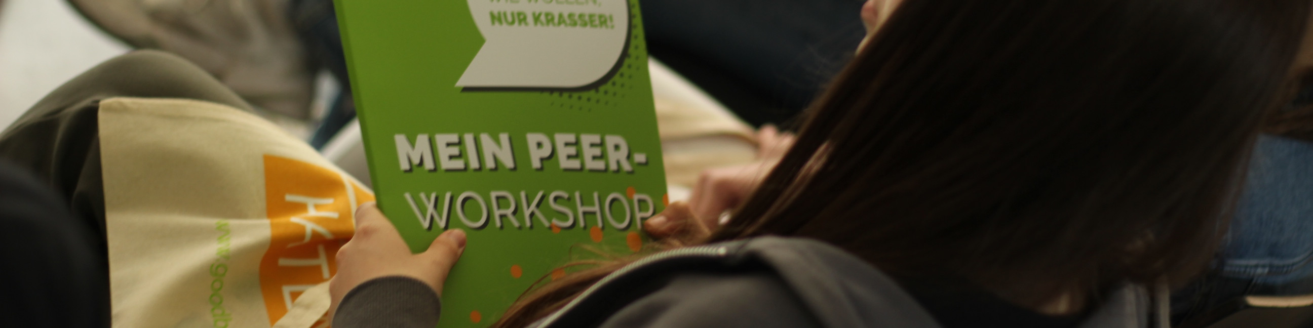 KI generiert: Das Bild zeigt eine Person, die ein grünes Schild mit der Aufschrift "MEIN PEER-WORKSHOP" und "NUR KRASSE!" hält. Im Vordergrund ist eine weitere Person mit einer Tüte zu sehen, auf der teilweise "RITA" zu lesen ist.