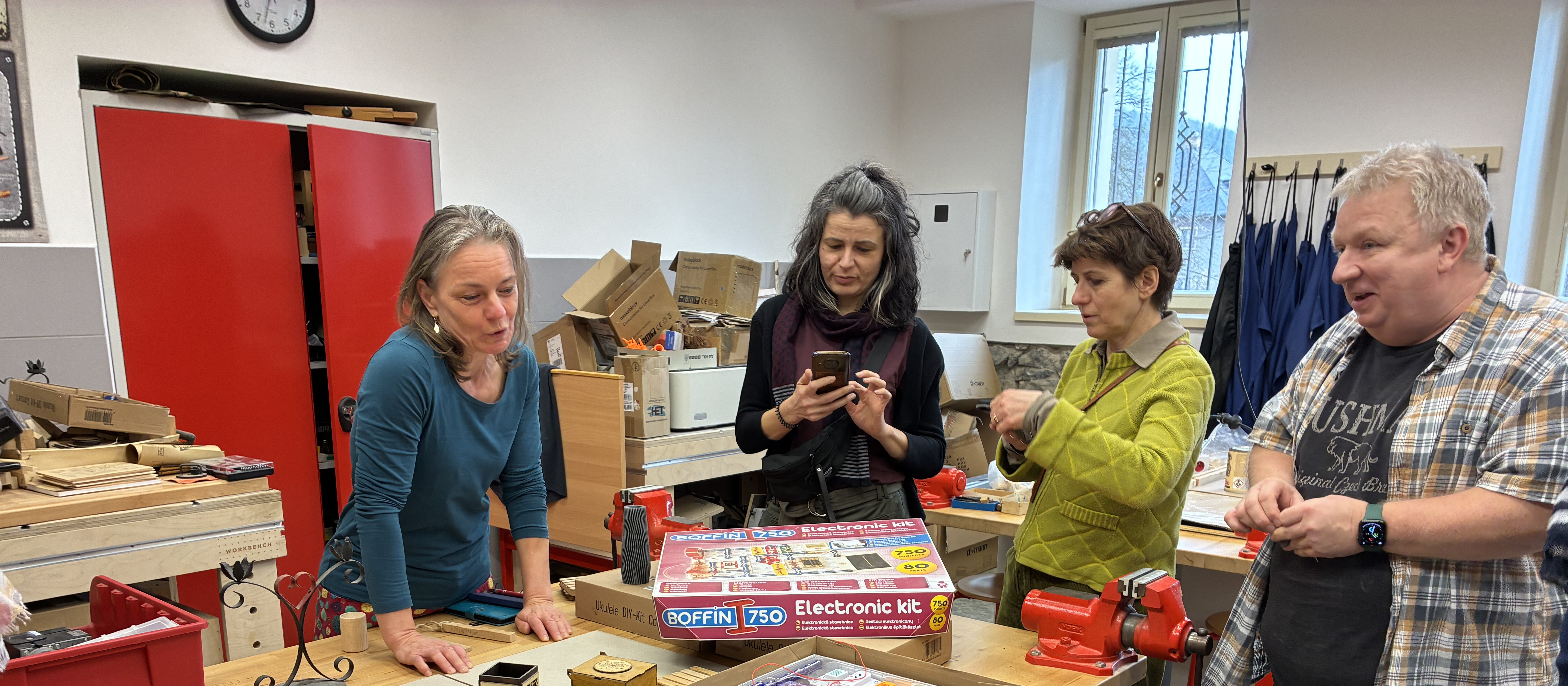 KI generiert: Das Bild zeigt vier Personen in einer Werkstatt, die sich um einen Tisch mit einem elektronischen Bausatz versammelt haben. Der Hauptinhalt des Bildes ist eine Gruppe von Menschen, die gemeinsam an einem Projekt arbeiten oder sich darüber austauschen.
