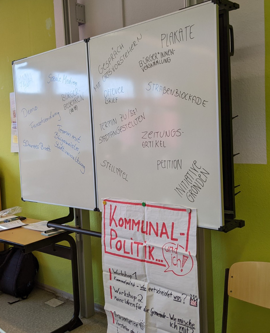 KI generiert: Das Bild zeigt ein Klassenzimmer mit einem Whiteboard, auf dem verschiedene Begriffe und Notizen zu einem Thema aus der Kommunalpolitik geschrieben stehen. Im Vordergrund hängt ein Plakat mit der Aufschrift "Kommunal-Politik... und ich?" sowie weiteren Informationen zu Workshops.