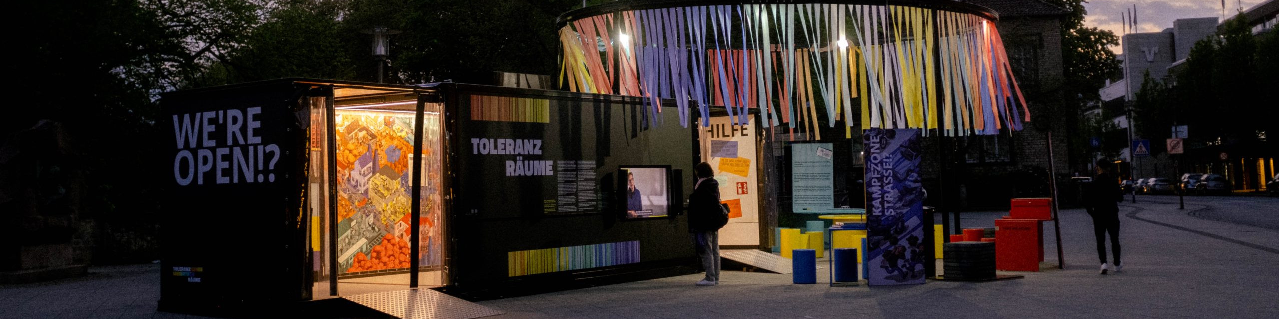 KI generiert: Das Bild zeigt eine abendliche Szene mit einem beleuchteten, bunten Pavillon auf einem Platz, der als "Toleranz Räume" beschriftet ist. Der Hauptinhalt des Bildes ist der Pavillon, der Kunstwerke und Informationen präsentiert und zum Betreten einlädt.