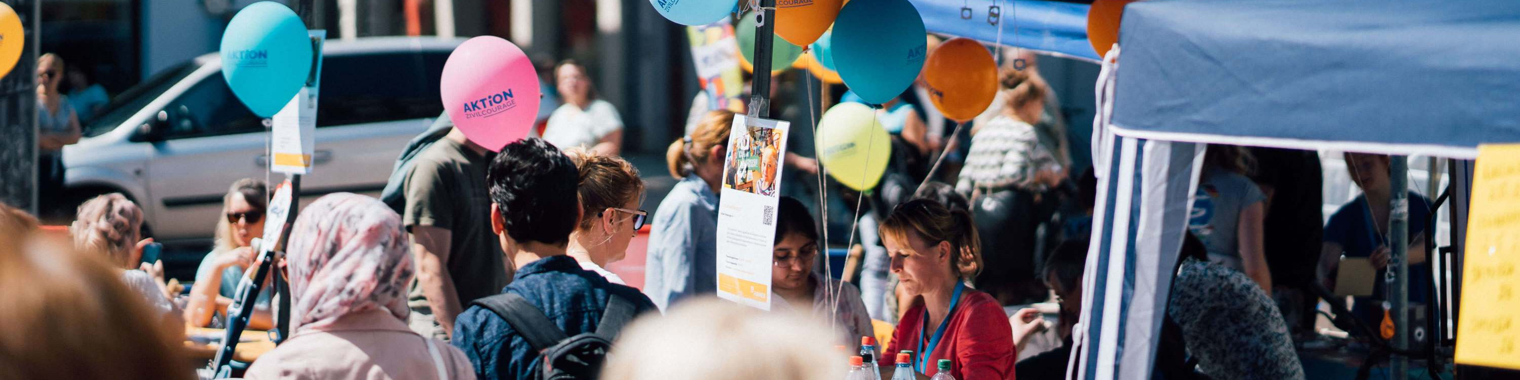 KI generiert: Das Bild zeigt eine belebte Straßenveranstaltung mit vielen Menschen, bunten Ballons und einem Informationsstand unter einem blauen Zelt. Im Vordergrund sind Personen in einer geselligen Atmosphäre, während im Hintergrund weitere Besucher und ein geparktes Auto zu sehen sind.