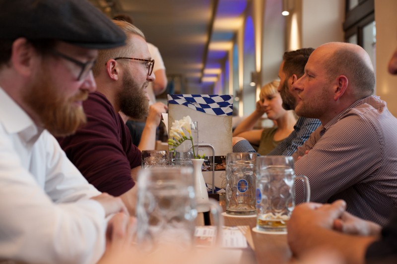 KI generiert: Das Bild zeigt eine Gruppe von Personen, die in einem geselligen Rahmen an einem Tisch sitzen und sich unterhalten. Im Vordergrund sind Bierkrüge und ein Tablett mit einer blau-weißen Karomuster-Serviette zu sehen.