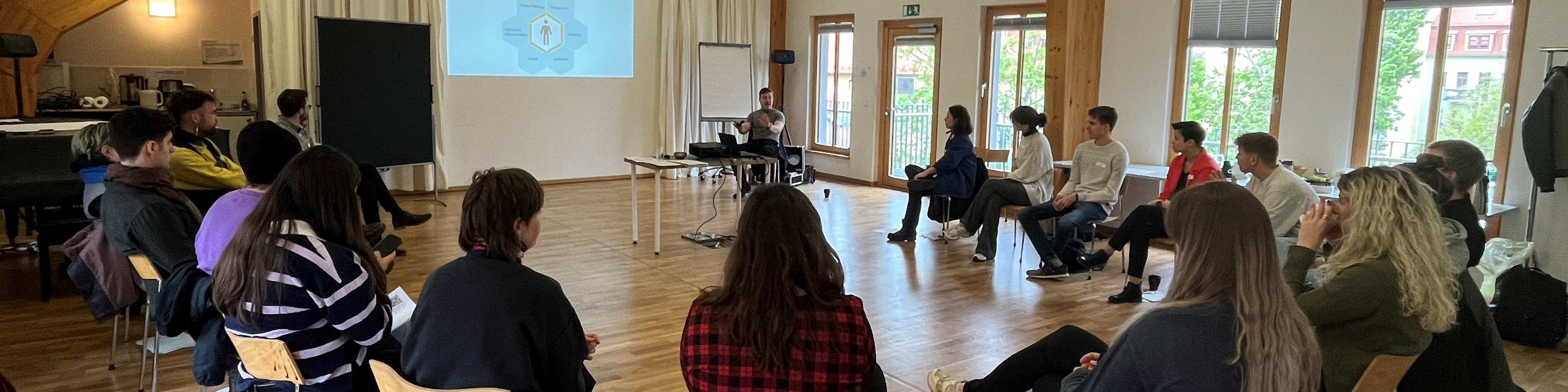 KI generiert: Das Bild zeigt eine Gruppe von Personen, die in einem hellen Raum mit Holzfußboden sitzen und einer Präsentation oder einem Vortrag folgen. Im Vordergrund sind Zuhörer zu sehen, während im Hintergrund eine Person vor einem Projektor steht und eine Präsentation hält.