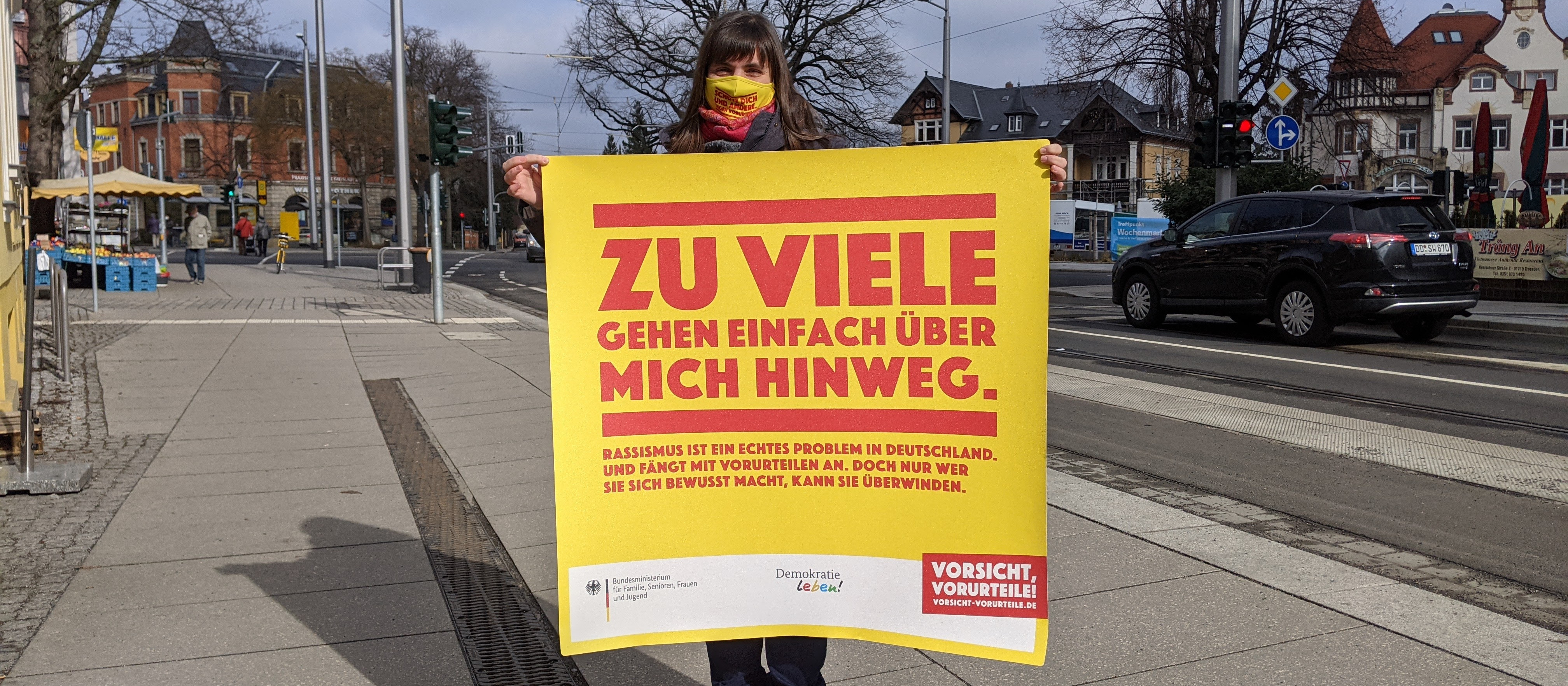 KI generiert: Das Bild zeigt eine Person, die auf einem Gehweg steht und ein gelbes Plakat mit roter Schrift hält, das auf Deutsch "Zu viele gehen einfach über mich hinweg" bedeutet. Im Hintergrund sind eine Straße, Gebäude und ein Auto zu sehen.