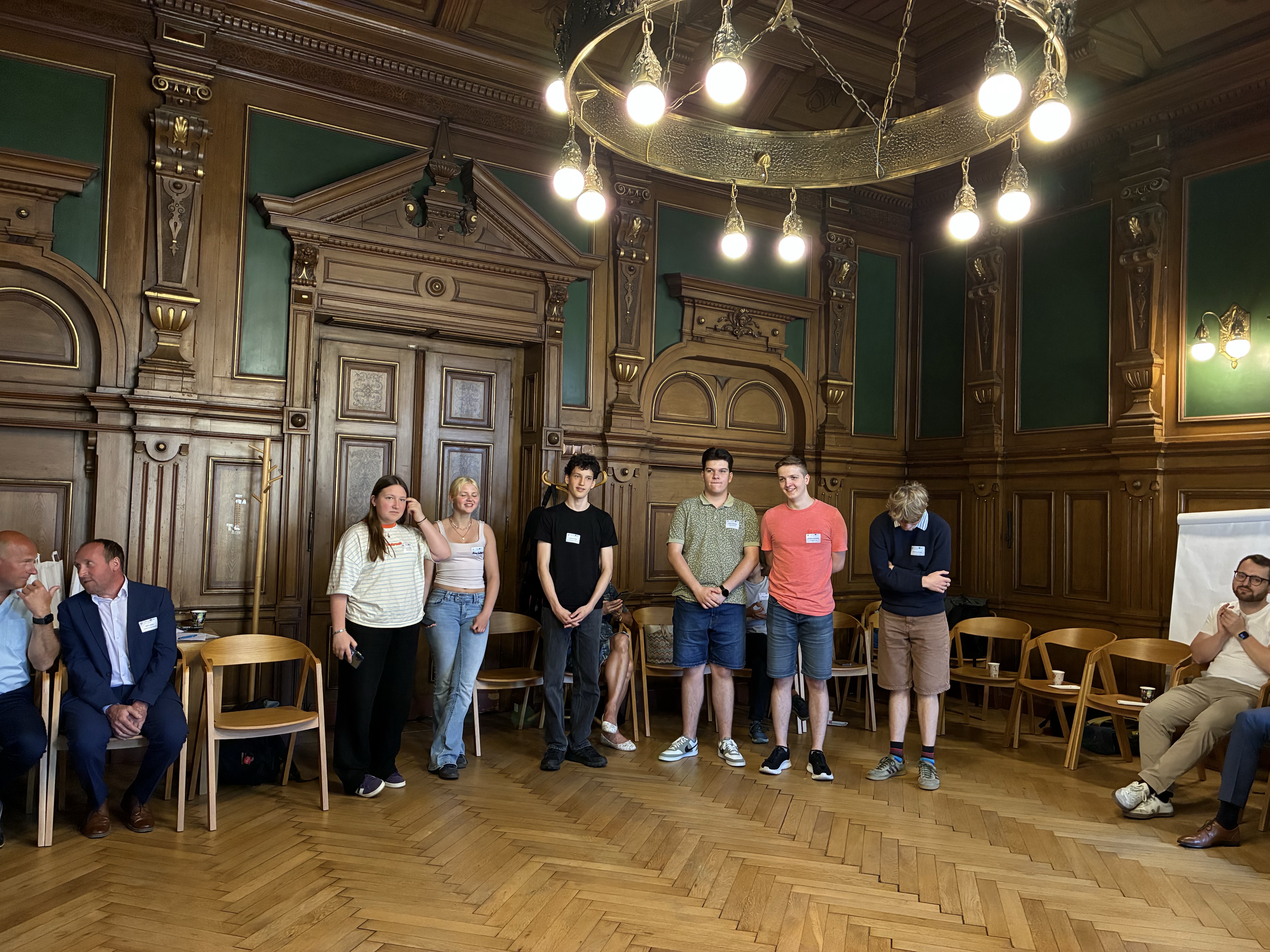 KI generiert: Das Bild zeigt eine Gruppe von Personen in einem historischen Raum mit Holzvertäfelungen und einem großen Kronleuchter. Die meisten Personen stehen in der Mitte des Raumes, während einige am Rand sitzen, und es scheint sich um eine Versammlung oder ein Meeting zu handeln.