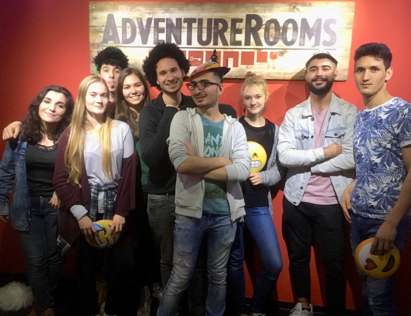 KI generiert: Das Bild zeigt eine Gruppe von neun Personen, die vor einem Schild mit der Aufschrift "ADVENTUREROOMS" posieren. Die Personen wirken fröhlich und halten teilweise Accessoires wie eine Tasche und ein Emoji-Kissen.