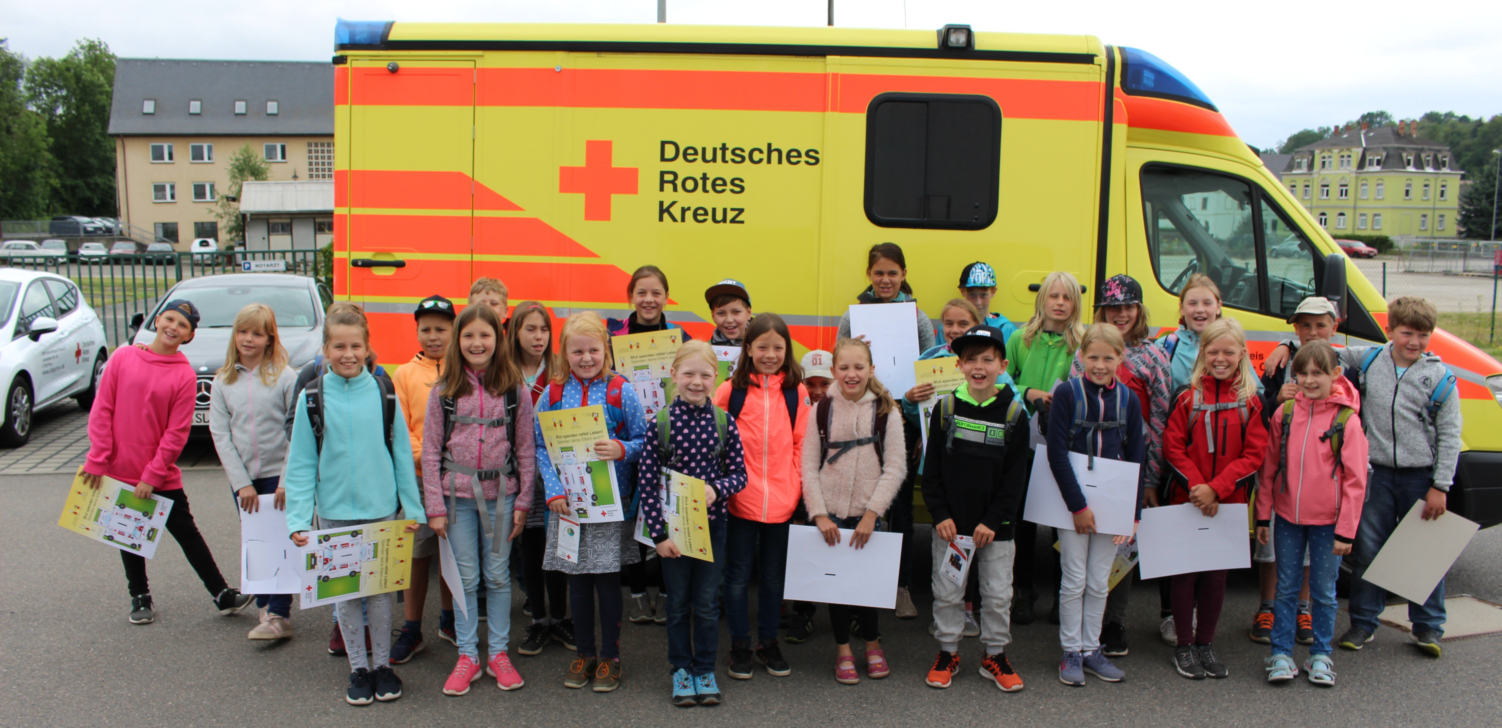 KI generiert: Das Bild zeigt eine Gruppe von Kindern, die stolz ihre Zertifikate vor einem Rettungswagen des Deutschen Roten Kreuzes präsentieren. Der Hauptinhalt des Bildes ist die feierliche Übergabe der Zertifikate, möglicherweise nach einem Erste-Hilfe-Kurs.