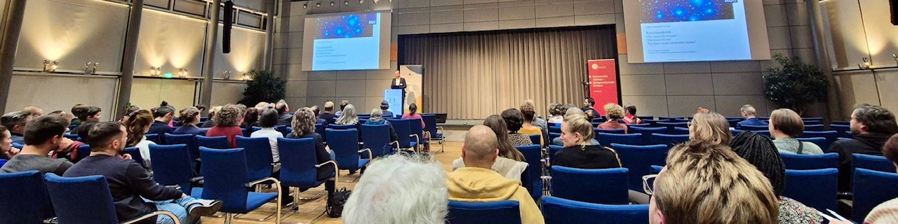 KI generiert: Das Bild zeigt eine Vortragsveranstaltung in einem großen, gut besuchten Hörsaal. Eine Person hält einen Vortrag auf der Bühne, während das Publikum in blauen Sitzen zuhört und zwei Projektionsleinwände mit Präsentationen im Hintergrund sichtbar sind.