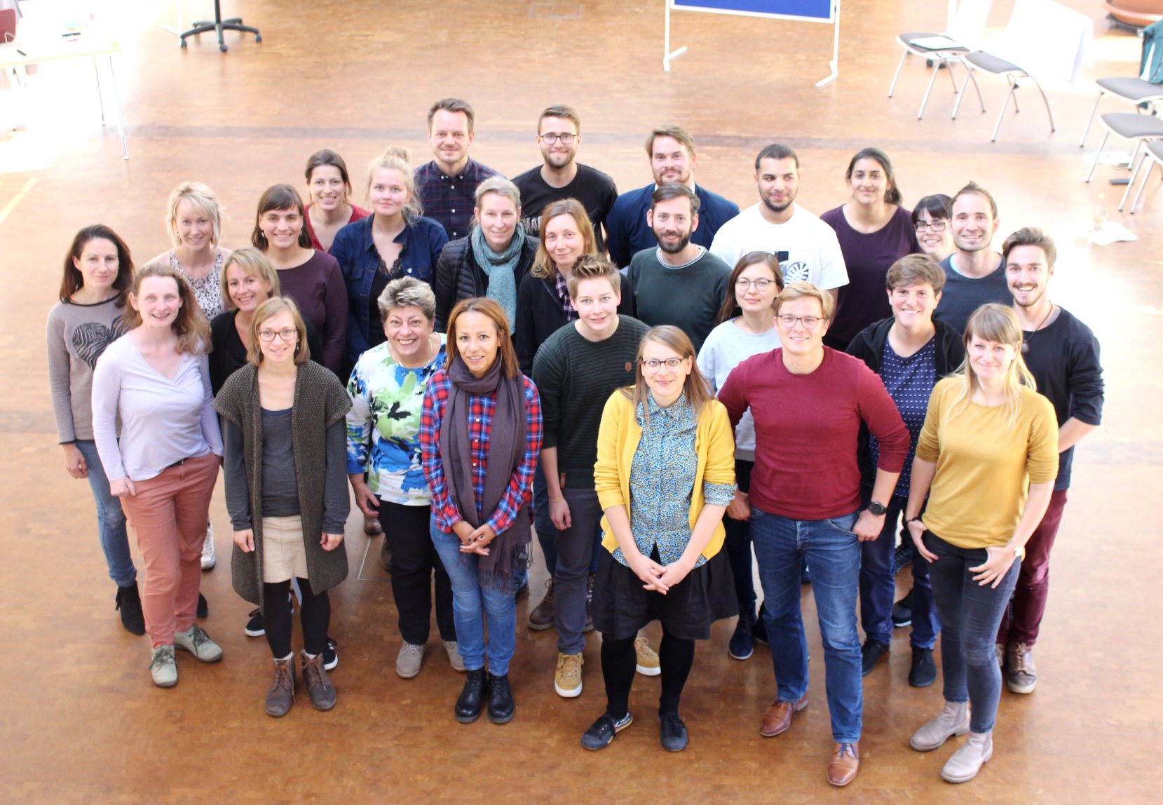 KI generiert: Das Bild zeigt eine Gruppe von etwa 25 Personen, die in einem hellen Innenraum mit Holzfußboden und einigen Stühlen im Hintergrund posieren. Der Hauptinhalt des Bildes ist die Gruppenfotografie, die eine vielfältige Gruppe von Menschen in lockerer, freundlicher Atmosphäre zeigt.