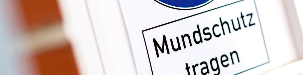 KI generiert: Das Bild zeigt ein Schild mit der Aufschrift "Mundschutz tragen". Der Hauptinhalt des Bildes ist die Aufforderung, einen Mundschutz zu tragen, was auf eine Maßnahme zur Infektionsprävention hinweist.