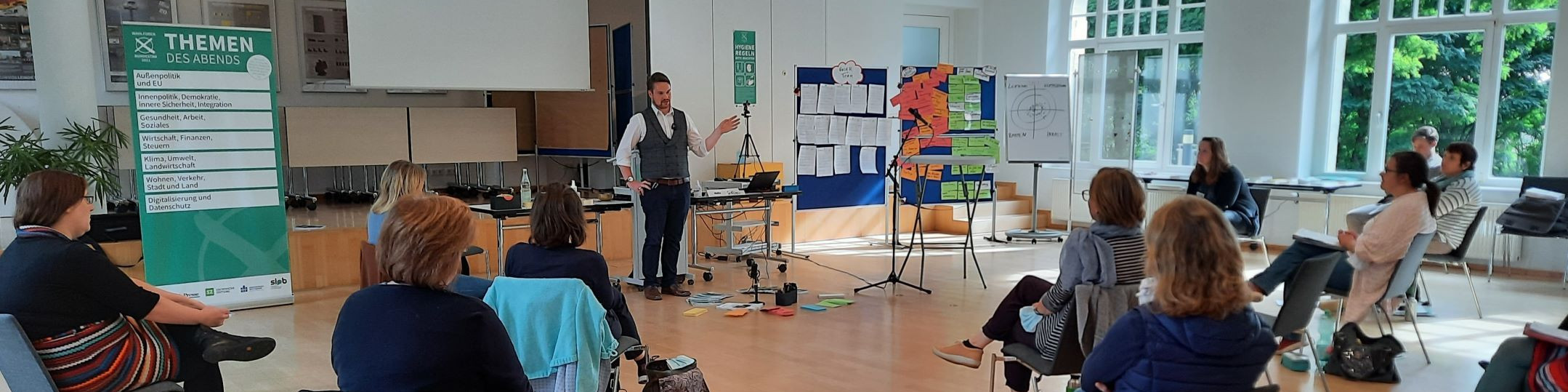 KI generiert: Das Bild zeigt eine Gruppe von Personen in einem hellen Raum, die an einer Präsentation oder einem Workshop teilnehmen. Ein Mann steht vorne und präsentiert Inhalte, während die Zuhörer auf Stühlen sitzen und zuhören.