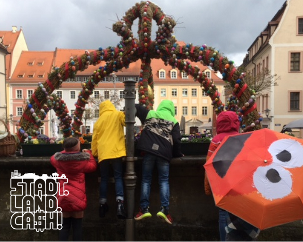 KI generiert: Das Bild zeigt eine belebte Straßenparade mit vielen Menschen, die sich um einen geschmückten Wagen mit einem großen, verzierten Kranz versammelt haben. Im Hintergrund sind historische Gebäude zu sehen, was auf eine traditionelle Feier oder einen Festumzug in einer Altstadt hindeutet.