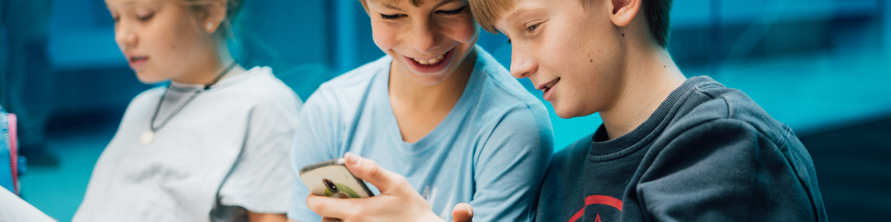 KI generiert: Das Bild zeigt drei Jugendliche, die gemeinsam auf ein Smartphone schauen und dabei lächeln. Der Hauptinhalt des Bildes ist die Interaktion und das gemeinsame Interesse der Jugendlichen an dem Inhalt des Smartphones.