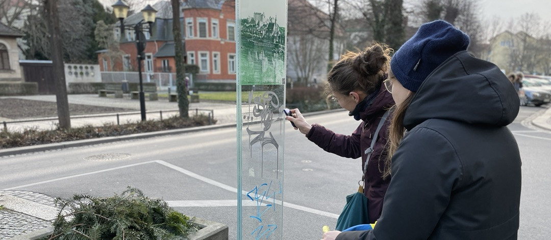 KI generiert: Das Bild zeigt zwei Personen, die an einer Glasfläche stehen und diese mit einem Werkzeug bearbeiten. Im Hintergrund sind eine Straße, Gebäude und Bäume zu sehen.