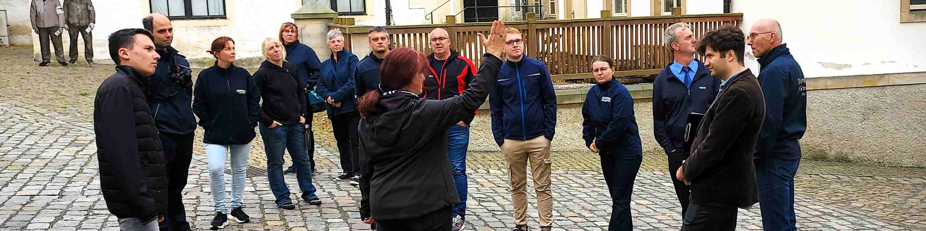 KI generiert: Das Bild zeigt eine Gruppe von Personen, die im Freien auf einem gepflasterten Platz stehen und sich zu unterhalten scheinen. Eine Person in der Mitte hebt die Hand, möglicherweise um eine Frage zu stellen oder eine Diskussion zu leiten.