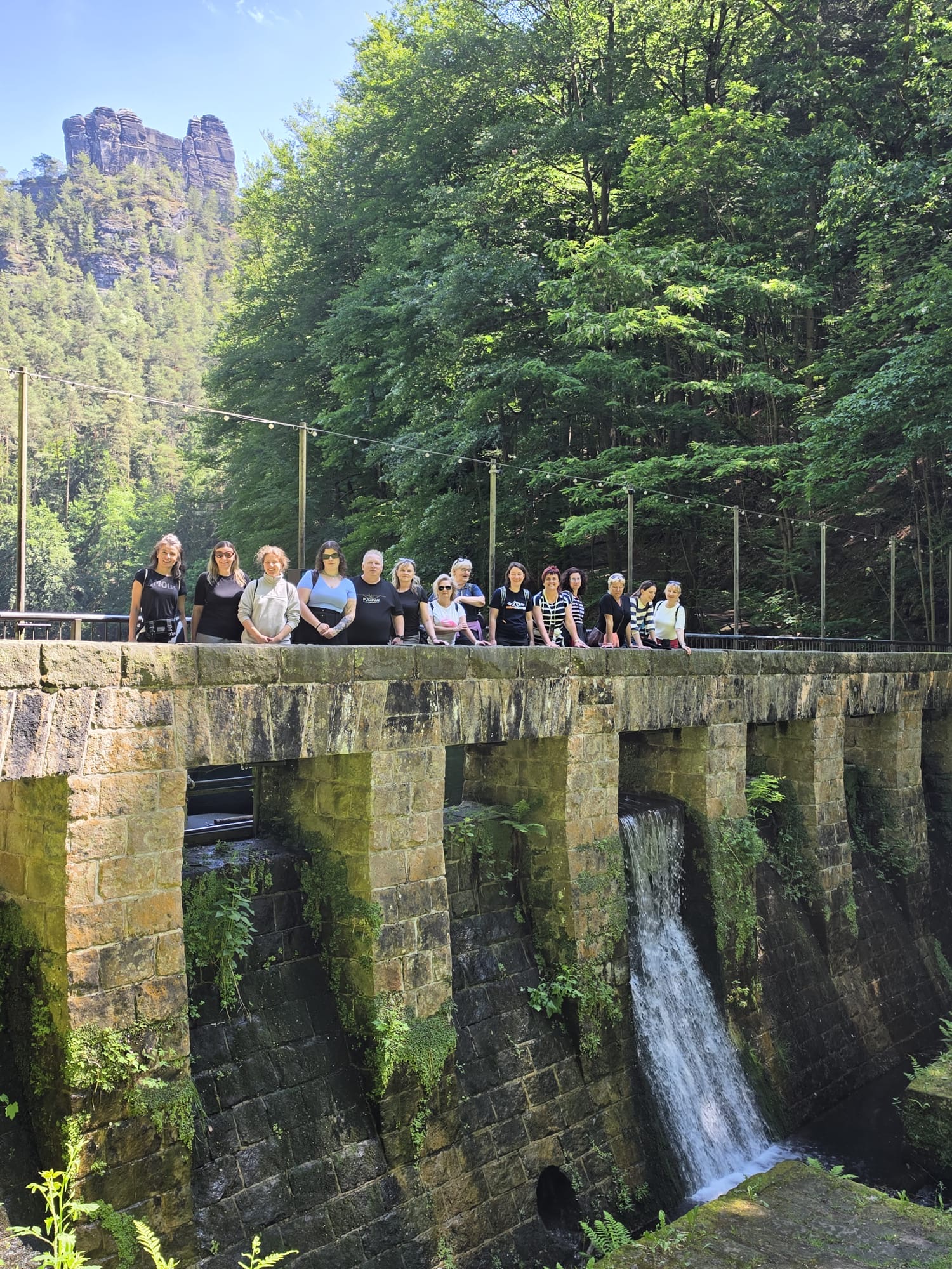 KI generiert: Das Bild zeigt eine Gruppe von Personen, die auf einer alten Steinbrücke über einem kleinen Wasserfall in einem bewaldeten Gebiet stehen. Im Hintergrund sind hohe Felsen und dichter Wald zu sehen.