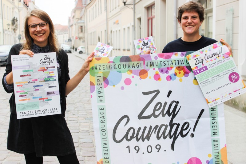KI generiert: Das Bild zeigt zwei Personen, die auf einer Straße stehen und Plakate sowie Flyer für den "Tag der Zivilcourage" am 19. September halten. Der Hauptinhalt des Bildes ist die Bewerbung dieser Veranstaltung, die sich für Zivilcourage einsetzt.