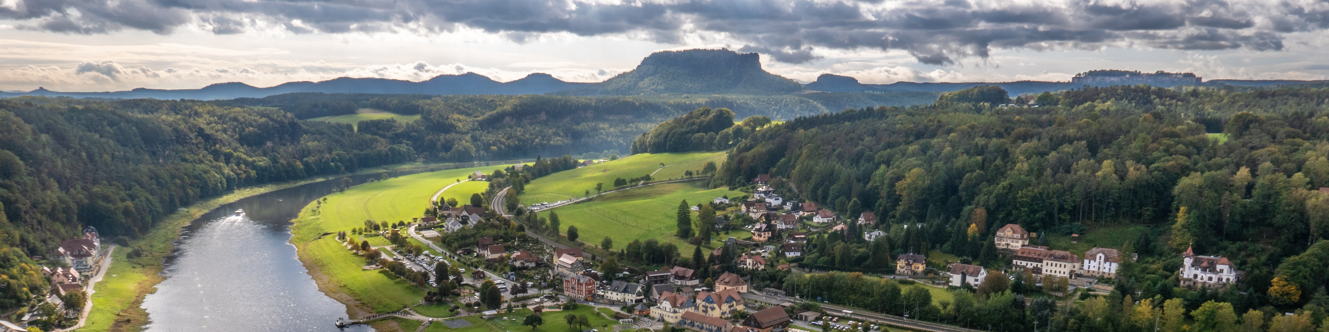 KI generiert: Das Bild zeigt eine malerische Landschaft mit einem Fluss, der sich durch eine grüne Talsohle schlängelt, umgeben von bewaldeten Hügeln und einem kleinen Dorf. Im Hintergrund ragen markante Felsformationen und Berge in den bewölkten Himmel.