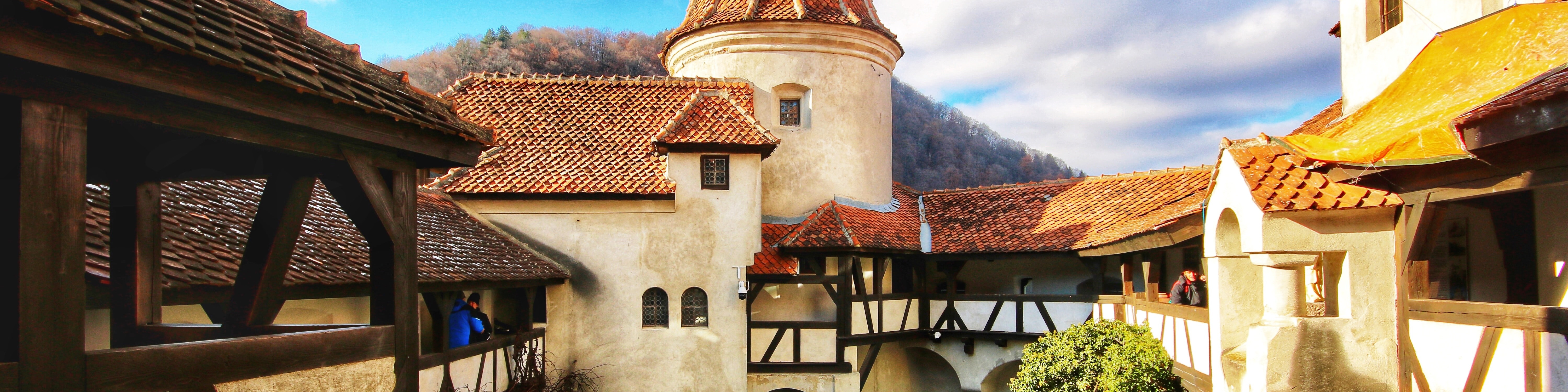 KI generiert: Das Bild zeigt eine malerische Burg mit roten Ziegeldächern und einem runden Turm, umgeben von einer bergigen Landschaft. Im Vordergrund sind Holzkonstruktionen und ein Innenhof zu sehen, in dem sich einige Personen aufhalten.