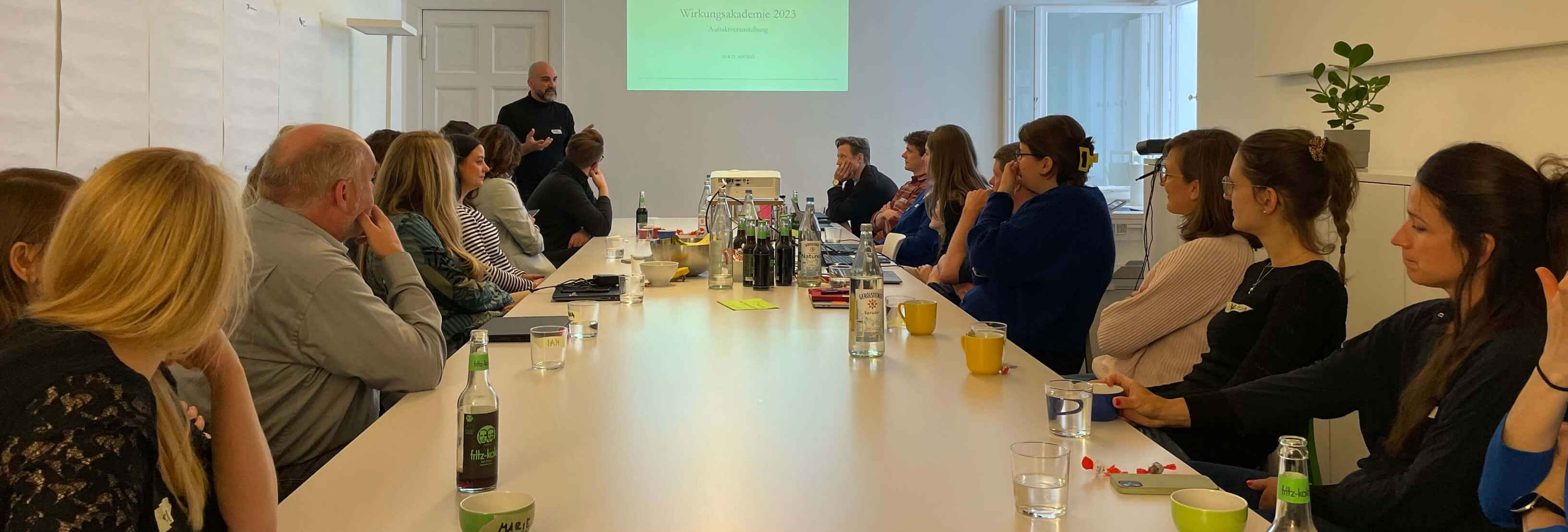KI generiert: Das Bild zeigt eine Gruppe von Personen, die an einem langen Tisch in einem hellen Raum sitzen und sich unterhalten. Im Hintergrund steht eine Person, die eine Präsentation hält, während eine Leinwand mit dem Titel "Willkommensworkshop 2017" zu sehen ist.