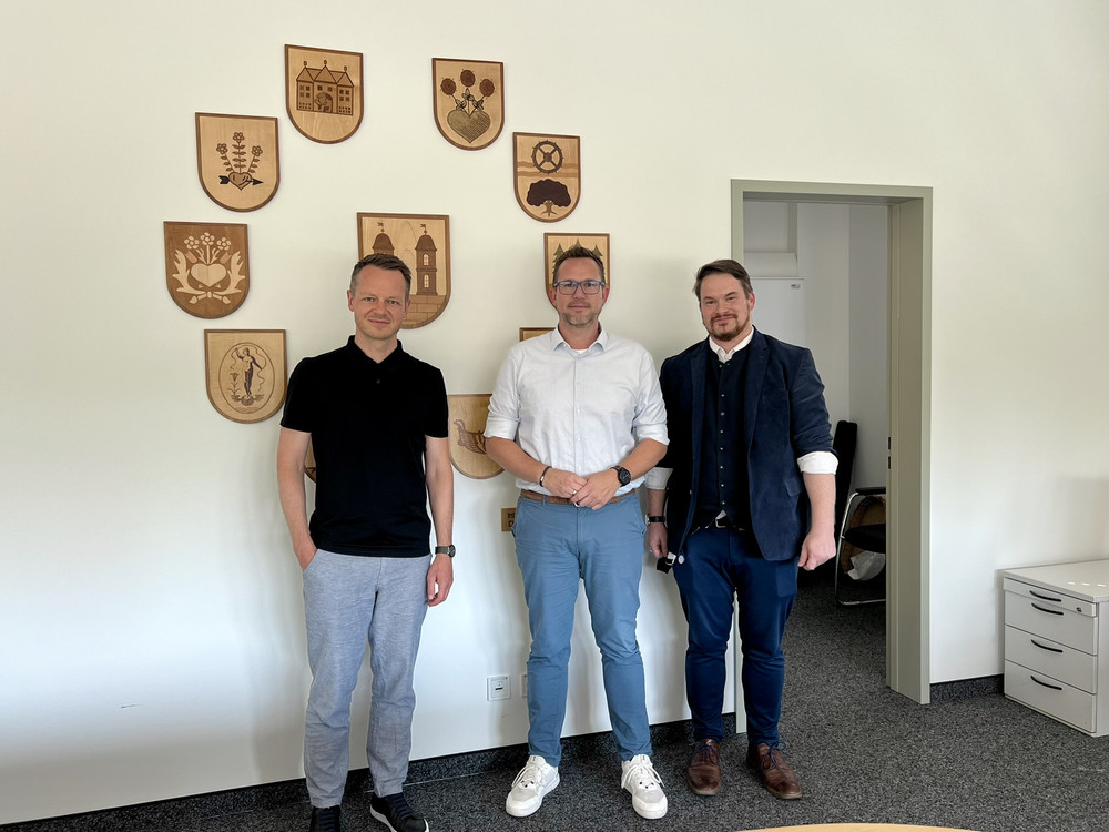 KI generiert: Das Bild zeigt drei Männer in einem Raum mit mehreren Holzschildern an der Wand, die verschiedene Symbole und Embleme darstellen. Die Personen stehen nebeneinander und posieren für das Foto.
