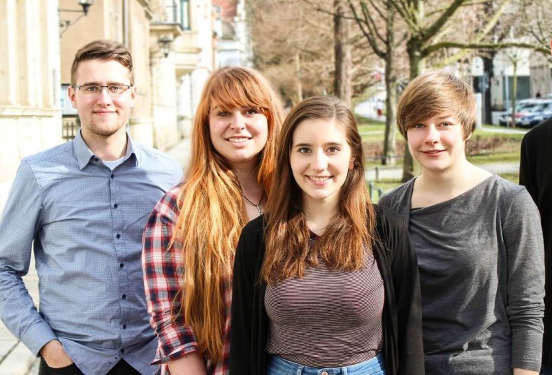KI generiert: Das Bild zeigt vier lächelnde junge Erwachsene, die eng beieinanderstehen und in die Kamera schauen. Sie stehen im Freien vor einem Gebäude mit Säulen und Bäumen im Hintergrund.