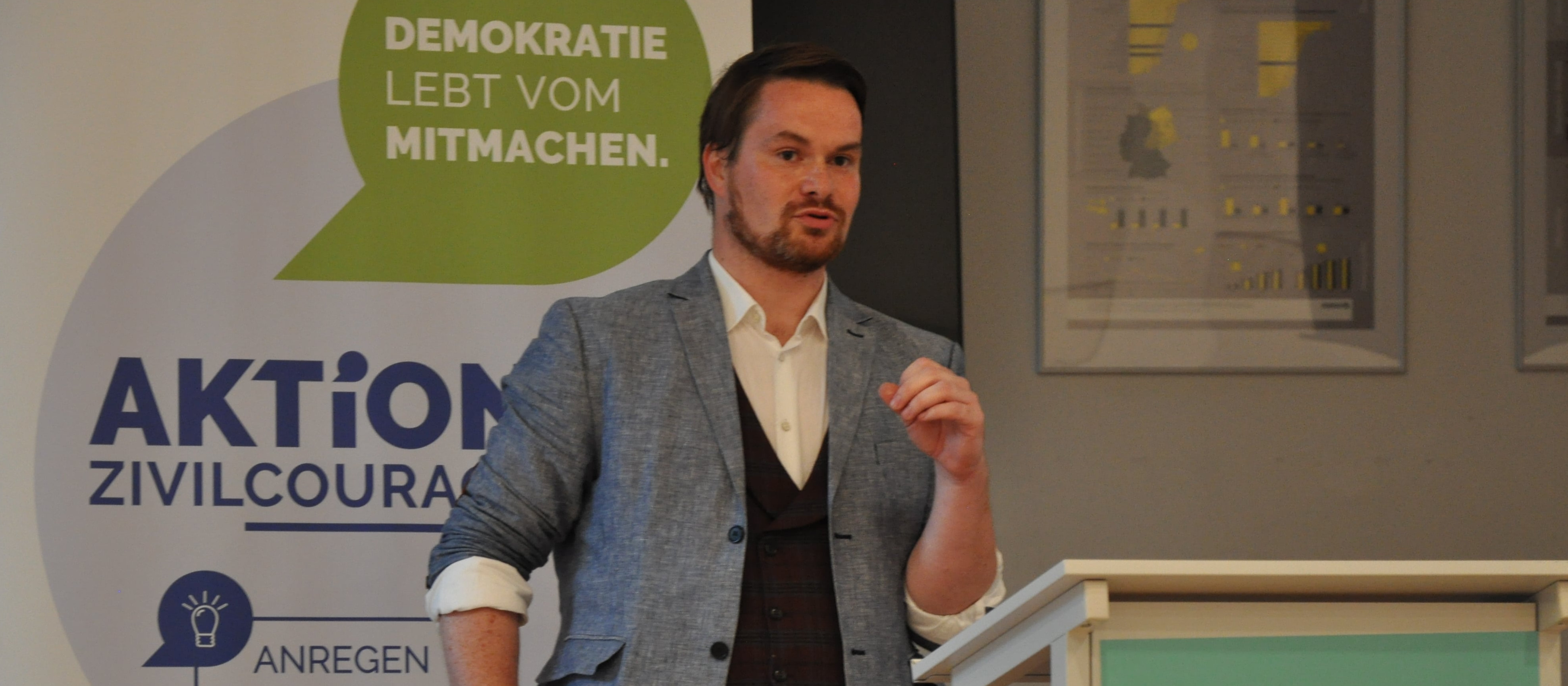 KI generiert: Das Bild zeigt einen Mann, der vor einem Podium steht und eine Rede hält. Im Hintergrund ist ein Banner mit der Aufschrift "AKTION ZIVILCOURAGE" und "DEMOKRATIE LEBT VOM MITMACHEN." zu sehen.