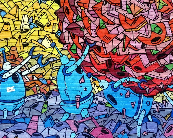 KI generiert: Das Bild zeigt eine bunte Graffiti-Wand mit verschiedenen cartoonartigen Robotern und abstrakten Formen. Der Hauptinhalt sind die beiden prominenten, blau gefärbten Roboterfiguren in der Mitte des Bildes.