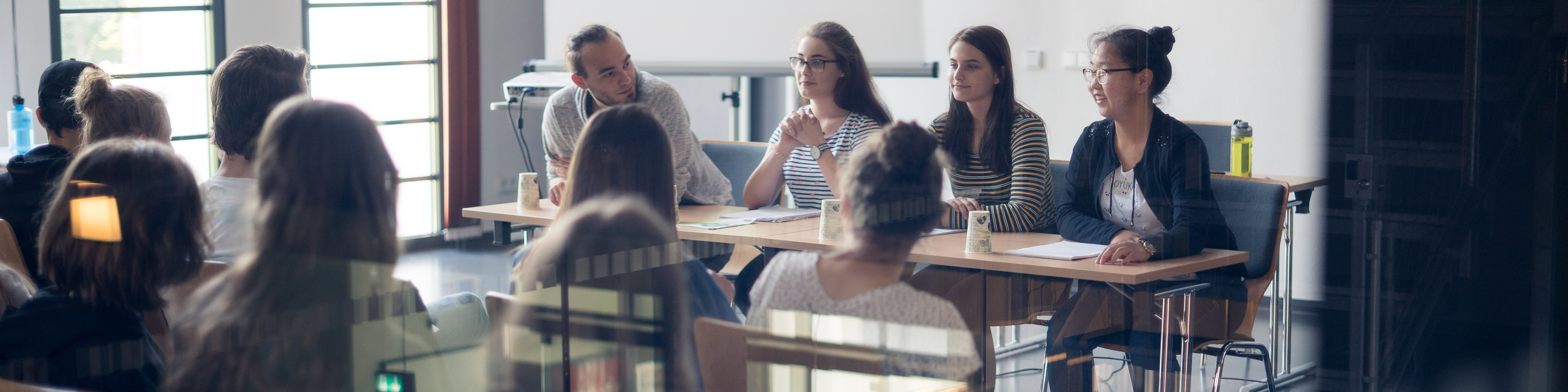 KI generiert: Das Bild zeigt eine Gruppe von Personen, die in einem hellen Raum an Tischen sitzen und sich in einer Diskussion oder einem Meeting befinden. Der Hauptinhalt des Bildes ist eine Besprechung oder ein Workshop in einem professionellen oder akademischen Umfeld.