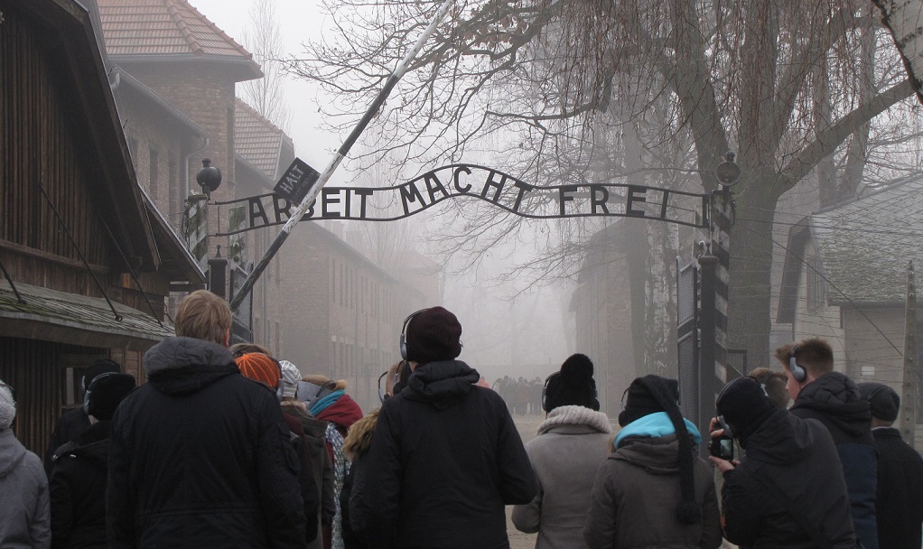 KI generiert: Das Bild zeigt eine Gruppe von Menschen, die durch das Tor mit der Aufschrift "Arbeit Macht Frei" am Eingang des ehemaligen Konzentrationslagers Auschwitz geht. Der Hauptinhalt des Bildes ist die symbolträchtige und historische Bedeutung des Ortes, der an die Gräueltaten des Holocaust erinnert.