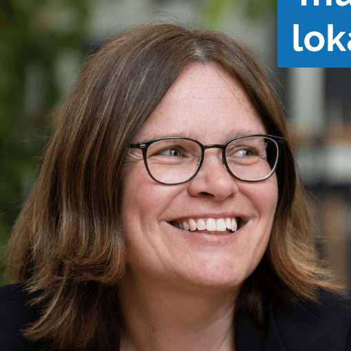 KI generiert: Das Bild zeigt eine lächelnde Frau mit Brille und braunem Haar, die in einem Innenraum mit Pflanzen im Hintergrund steht. Im oberen rechten Bereich des Bildes ist ein blaues Rechteck mit weißer Schrift zu sehen, das teilweise den Text "loka" enthält.