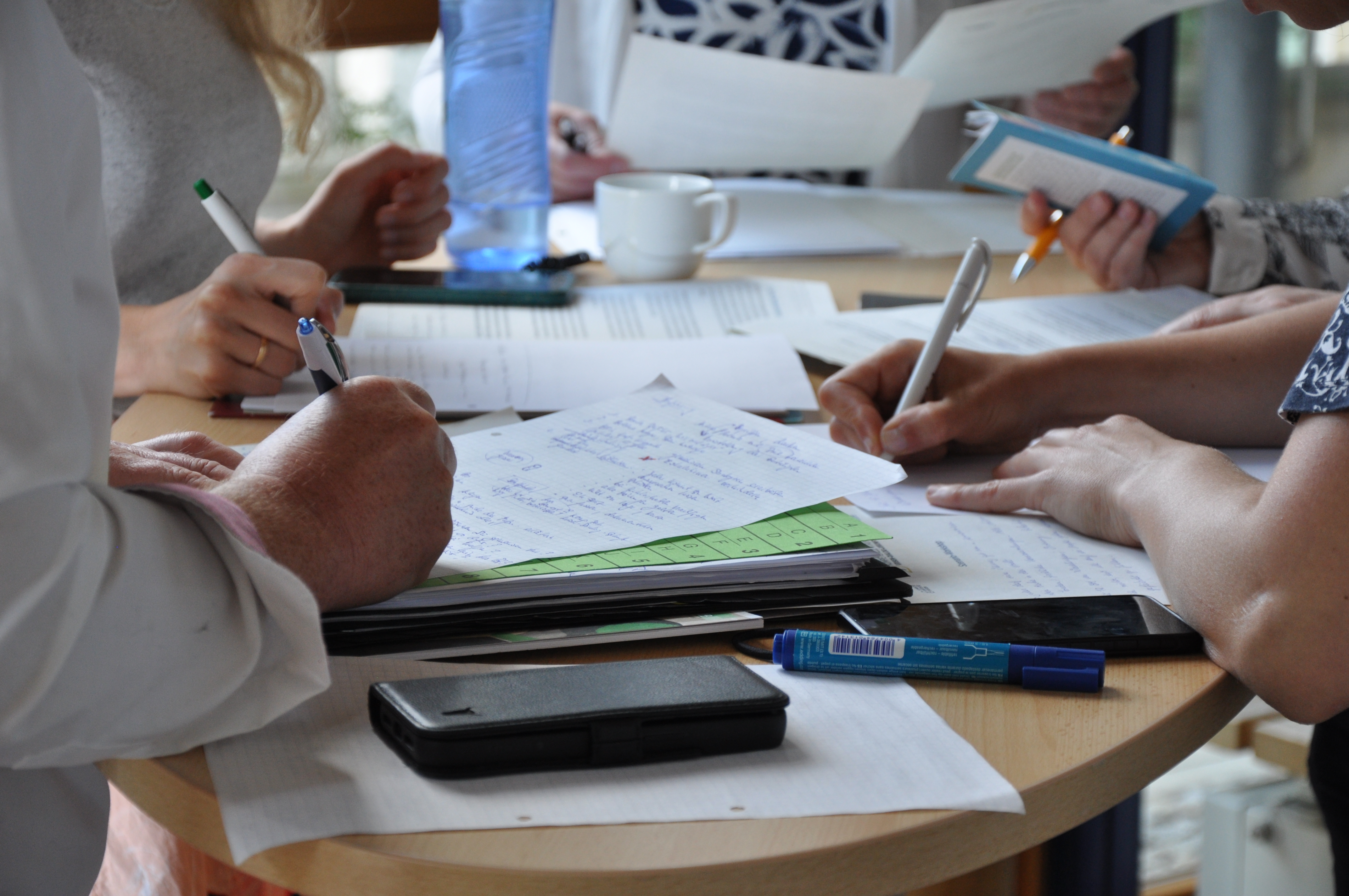 KI generiert: Das Bild zeigt eine Gruppe von Personen, die an einem runden Tisch sitzen und auf Papier schreiben oder zeichnen. Der Hauptinhalt des Bildes ist eine kreative oder Arbeitsbesprechung, bei der Notizen gemacht und Ideen ausgetauscht werden.