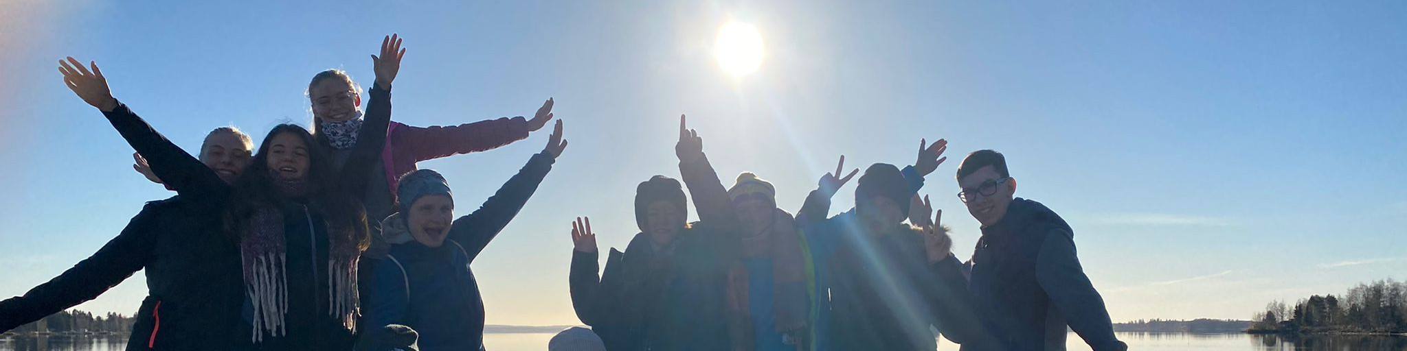 KI generiert: Das Bild zeigt eine Gruppe von Menschen, die draußen in der Natur stehen und fröhlich in die Kamera winken. Im Hintergrund ist ein See und ein klarer Himmel mit strahlendem Sonnenschein zu sehen.
