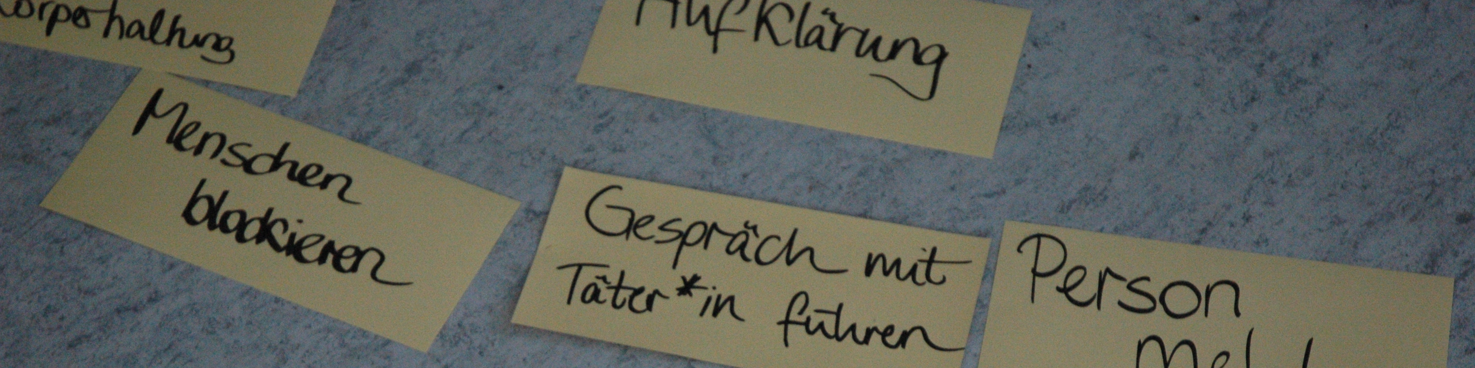 KI generiert: Das Bild zeigt mehrere gelbe Notizzettel auf einer grauen Oberfläche, auf denen handschriftliche Notizen in deutscher Sprache zu sehen sind. Die Notizen enthalten Begriffe wie "HWF-Klärung", "Menschen blockieren", "Gespräch mit Täter*in führen" und "Person mel".