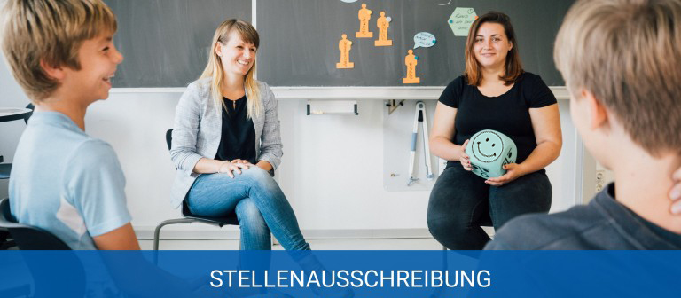 KI generiert: Das Bild zeigt eine Gruppenaktivität in einem Klassenzimmer, bei der zwei Erwachsene und zwei Kinder an einer Diskussion oder einem Spiel teilnehmen. Der Hauptinhalt des Bildes ist die Stellenausschreibung, die im unteren Bereich des Bildes hervorgehoben wird.