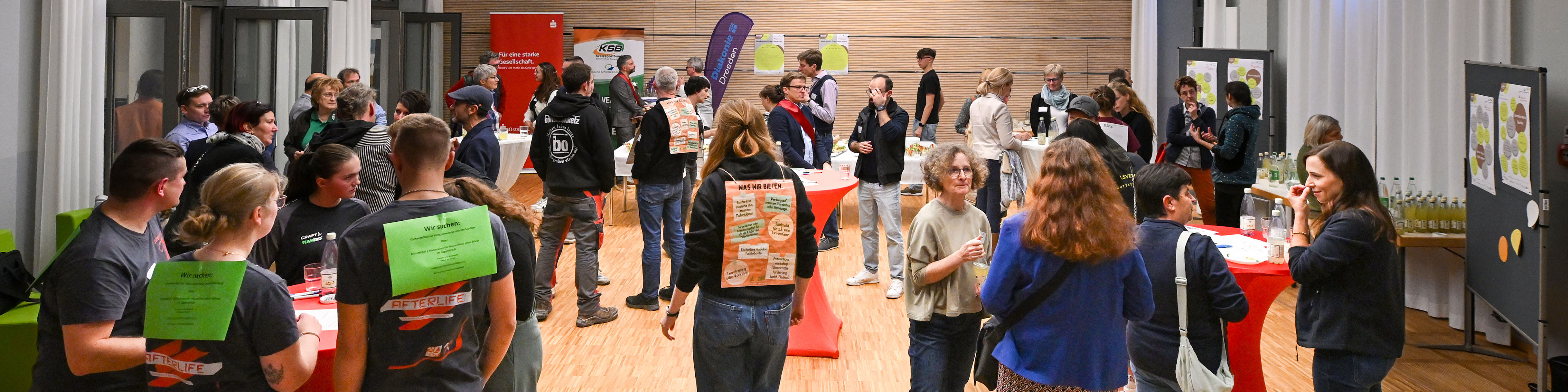 KI generiert: Das Bild zeigt eine belebte Indoor-Veranstaltung mit vielen Menschen, die sich an verschiedenen Ständen und Tischen unterhalten. Der Hauptinhalt des Bildes ist eine Netzwerk- oder Informationsveranstaltung, bei der die Teilnehmer in einer entspannten Atmosphäre interagieren.