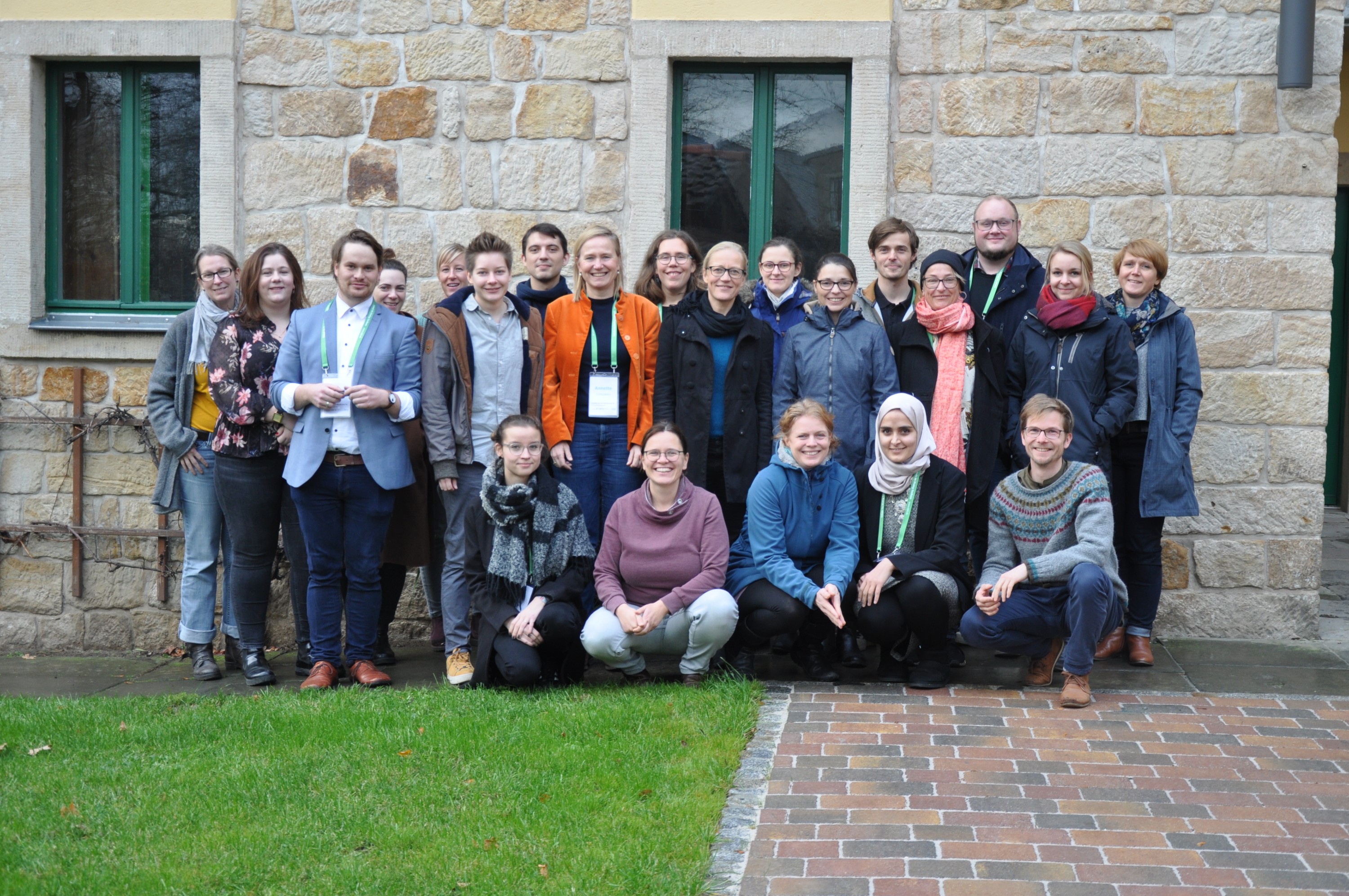 KI generiert: Das Bild zeigt eine Gruppe von Menschen, die vor einem steinernen Gebäude mit grünen Fenstern posieren. Der Hauptinhalt des Bildes ist die Gruppe von Personen, die gemeinsam ein Foto machen.