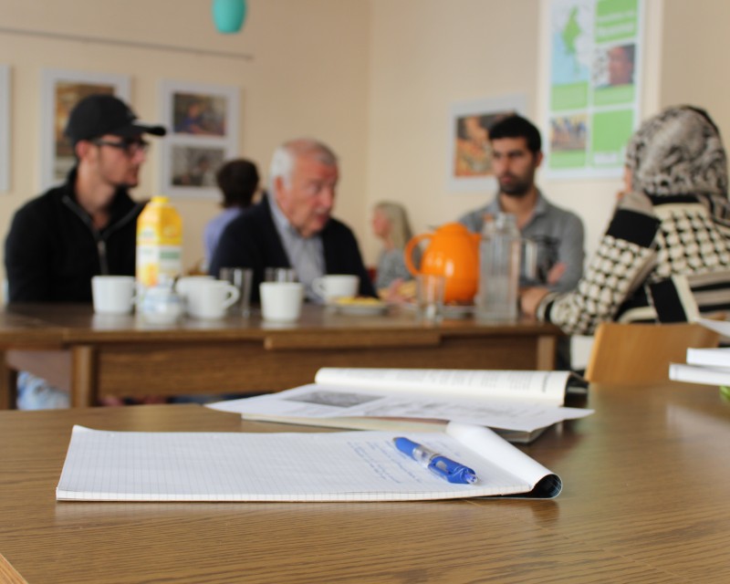 KI generiert: Das Bild zeigt eine Gruppe von Personen in einem Raum, die an Tischen sitzen und sich unterhalten, während Notizbücher und ein Stift im Vordergrund auf einem Tisch liegen. Der Hauptinhalt des Bildes ist eine informelle Zusammenkunft oder ein Treffen, möglicherweise ein Workshop oder eine Besprechung.