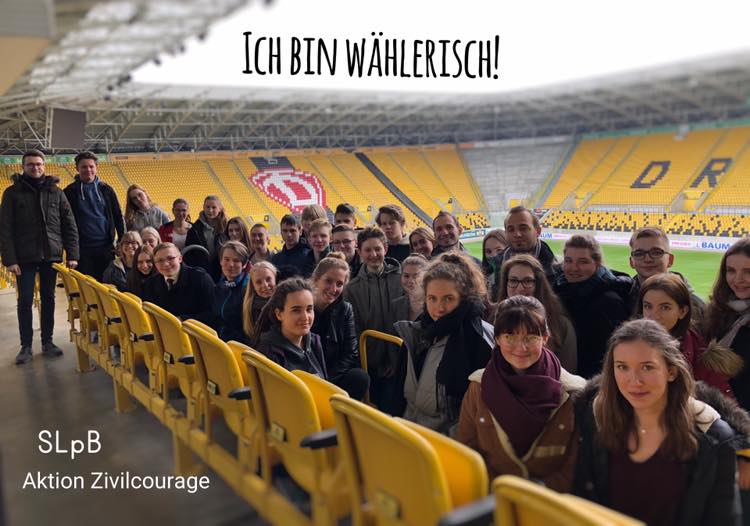 KI generiert: Das Bild zeigt eine Gruppe von Menschen, die in einem Stadion auf gelben Sitzen sitzen, mit dem Text "ICH BIN WÄHLERISCH!" und "SLpB Aktion Zivilcourage" darüber. Der Hauptinhalt des Bildes ist eine Kampagne zur Förderung von Zivilcourage und politischer Teilhabe.