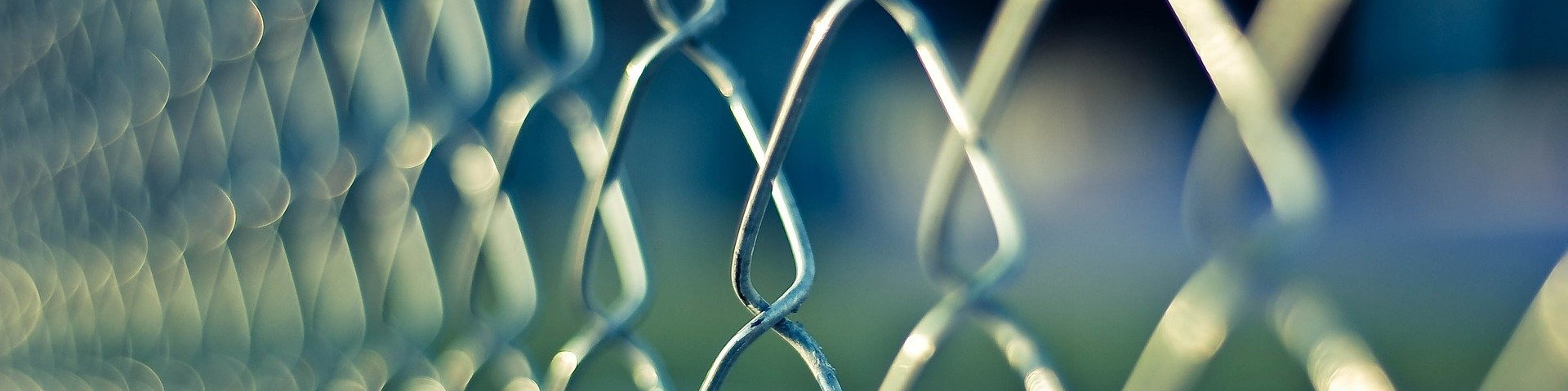 KI generiert: Das Bild zeigt eine Nahaufnahme eines Metallzauns mit rautenförmigen Maschen, der leicht unscharf im Hintergrund eine grüne Fläche erkennen lässt. Der Hauptinhalt des Bildes ist der Metallzaun.