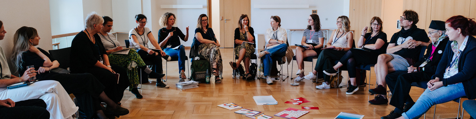 KI generiert: Das Bild zeigt eine Gruppe von Menschen, die in einem Kreis sitzen und sich in einer Diskussions- oder Arbeitsgruppe unterhalten. Der Hauptinhalt des Bildes ist eine interaktive Sitzung oder ein Workshop in einem hellen, holzgetäfelten Raum.