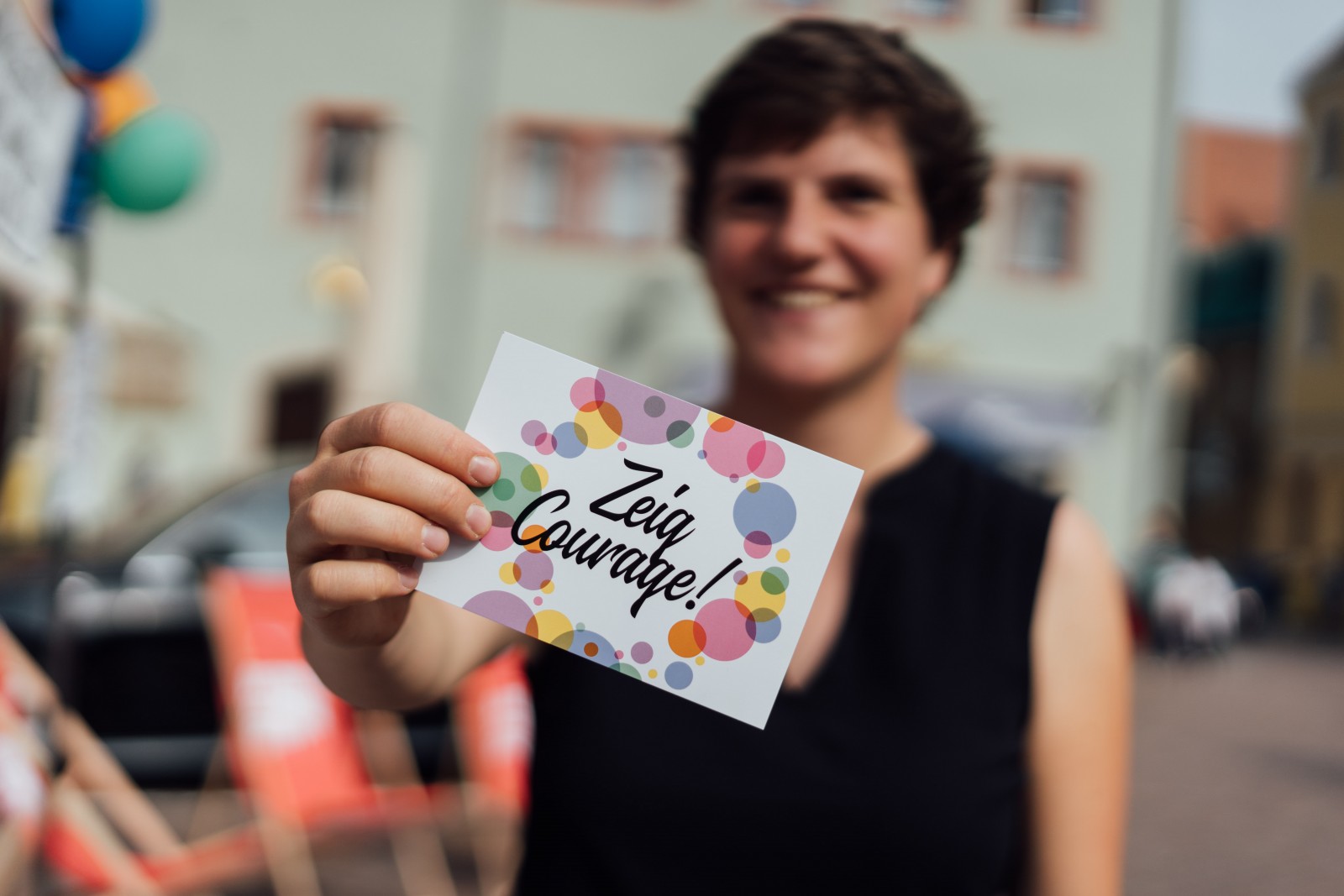 KI generiert: Das Bild zeigt eine lächelnde Person, die ein Kärtchen mit der Aufschrift "Zeig Courage!" in der Hand hält. Im Hintergrund sind bunte Ballons und eine Straßenumgebung zu erkennen.