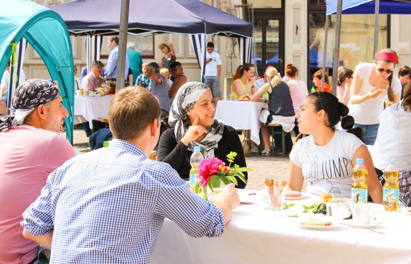 KI generiert: Das Bild zeigt eine Gruppe von Menschen, die an einem langen Tisch im Freien sitzen und sich unterhalten, während sie gemeinsam essen. Im Hintergrund sind weitere Tische mit Menschen und ein Zelt zu sehen, was auf eine gesellige Veranstaltung oder ein Fest hindeutet.
