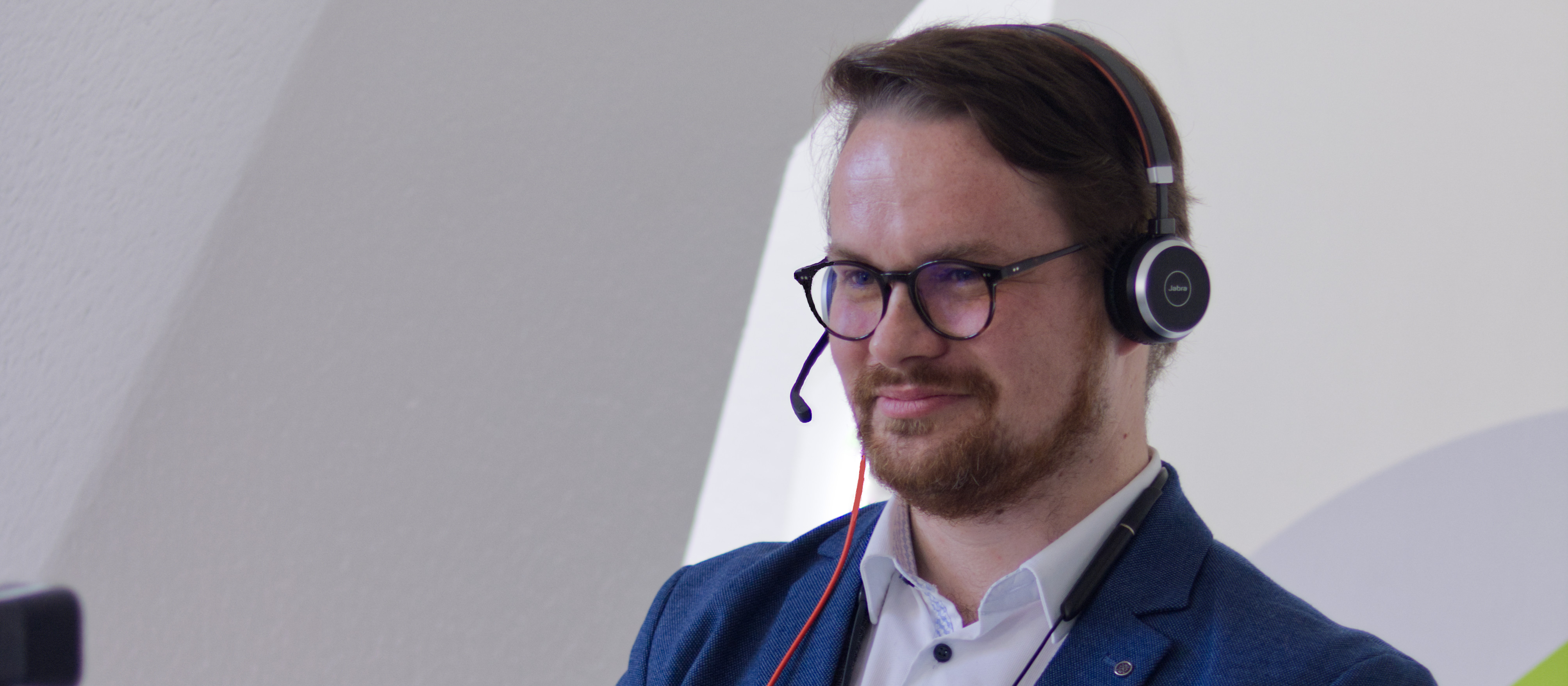 KI generiert: Das Bild zeigt einen Mann mit Brille und Kopfhörern, der einen Anzug trägt und in eine Kamera oder einen Bildschirm schaut. Der Hauptinhalt des Bildes ist der Mann, der möglicherweise an einer Videokonferenz oder einer Präsentation teilnimmt.
