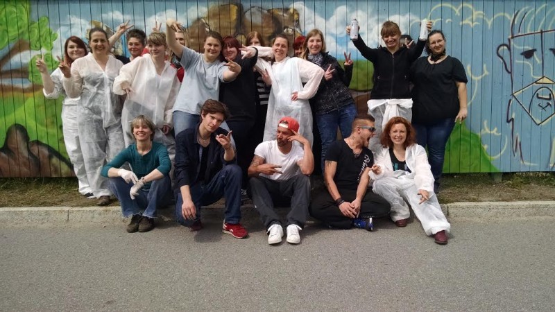 KI generiert: Das Bild zeigt eine Gruppe von Personen, die vor einer bunten Graffiti-Wand posieren. Viele von ihnen tragen weiße Schutzanzüge und einige halten Dosen, möglicherweise Farbdosen, in den Händen.