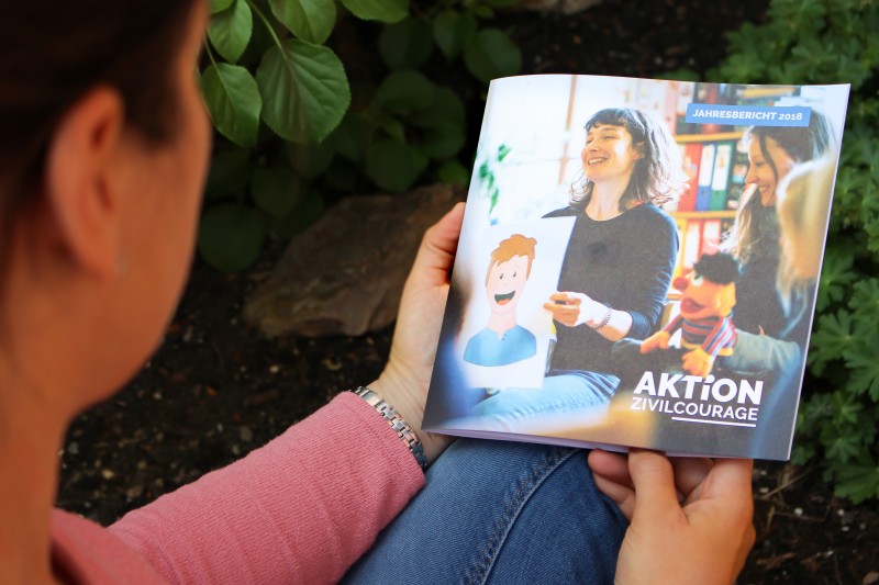 KI generiert: Das Bild zeigt eine Person, die im Freien sitzt und ein Jahresbericht 2018 der Organisation "Aktion Zivilcourage" liest. Der Bericht enthält ein Bild von Menschen, die sich engagieren, sowie Illustrationen, die Zivilcourage symbolisieren.