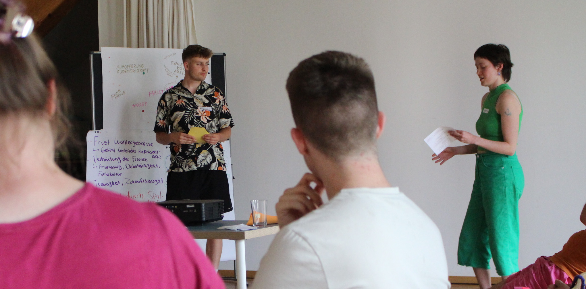 KI generiert: Das Bild zeigt eine kleine Gruppe von Personen in einem Raum, wobei eine Person vor einem Whiteboard steht und eine andere Person ihr gegenübersteht und Notizen zu machen scheint. Es handelt sich offenbar um eine Besprechung oder ein Seminar.