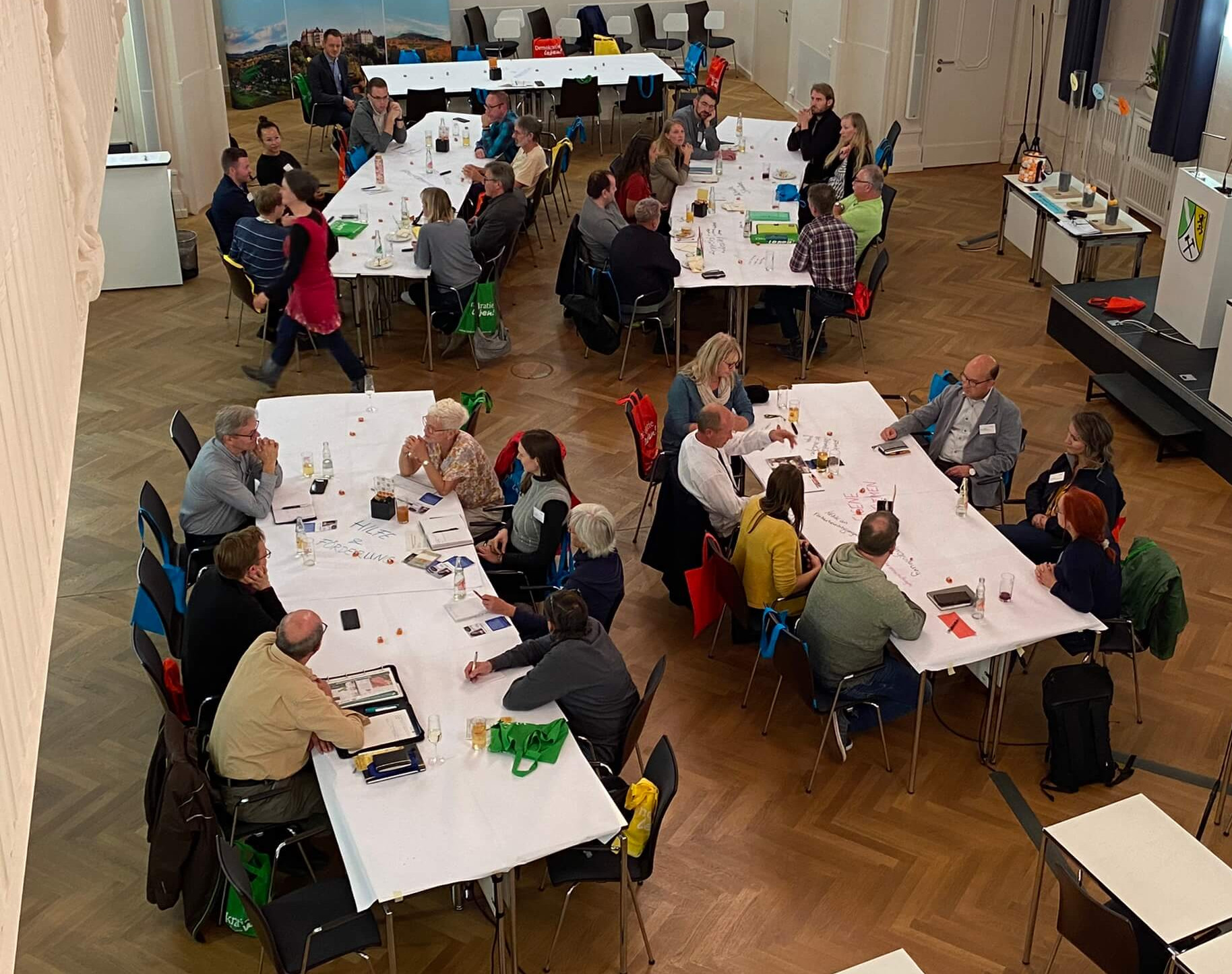 KI generiert: Das Bild zeigt eine Gruppe von Menschen, die in einem großen Raum an langen Tischen sitzen und sich unterhalten. Es scheint sich um eine informelle Zusammenkunft oder ein Meeting zu handeln, bei dem die Teilnehmer in kleinen Gruppen diskutieren.
