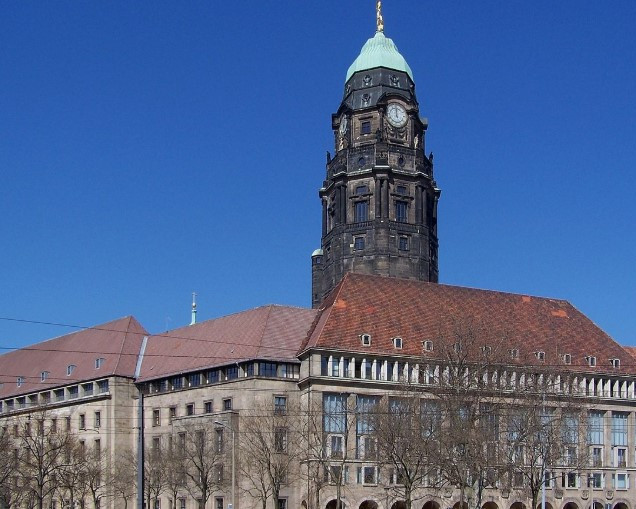 KI generiert: Das Bild zeigt den oberen Teil eines historischen Uhrturms mit einer grünen Kuppel und einer goldenen Spitze vor einem klaren blauen Himmel. Der Hauptinhalt des Bildes ist der Uhrturm.