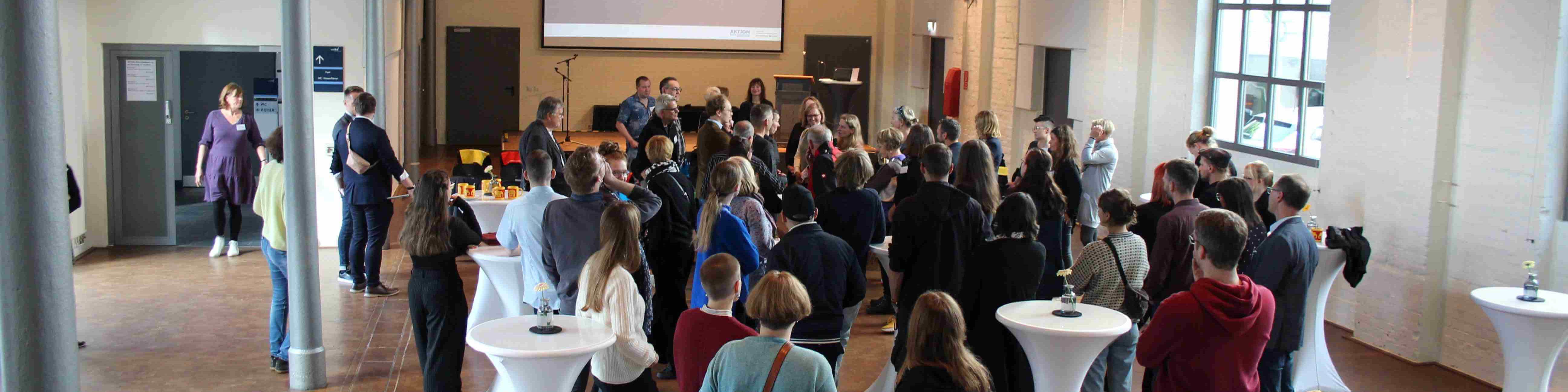 KI generiert: Das Bild zeigt eine Gruppe von Menschen, die in einem hellen, geräumigen Raum mit hohen Decken und großen Fenstern stehen und sich unterhalten. Im Raum gibt es mehrere Stehtische mit weißen Tischdecken, an denen einige Personen stehen, sowie eine kleine Bühne oder ein Podium im Hintergrund.
